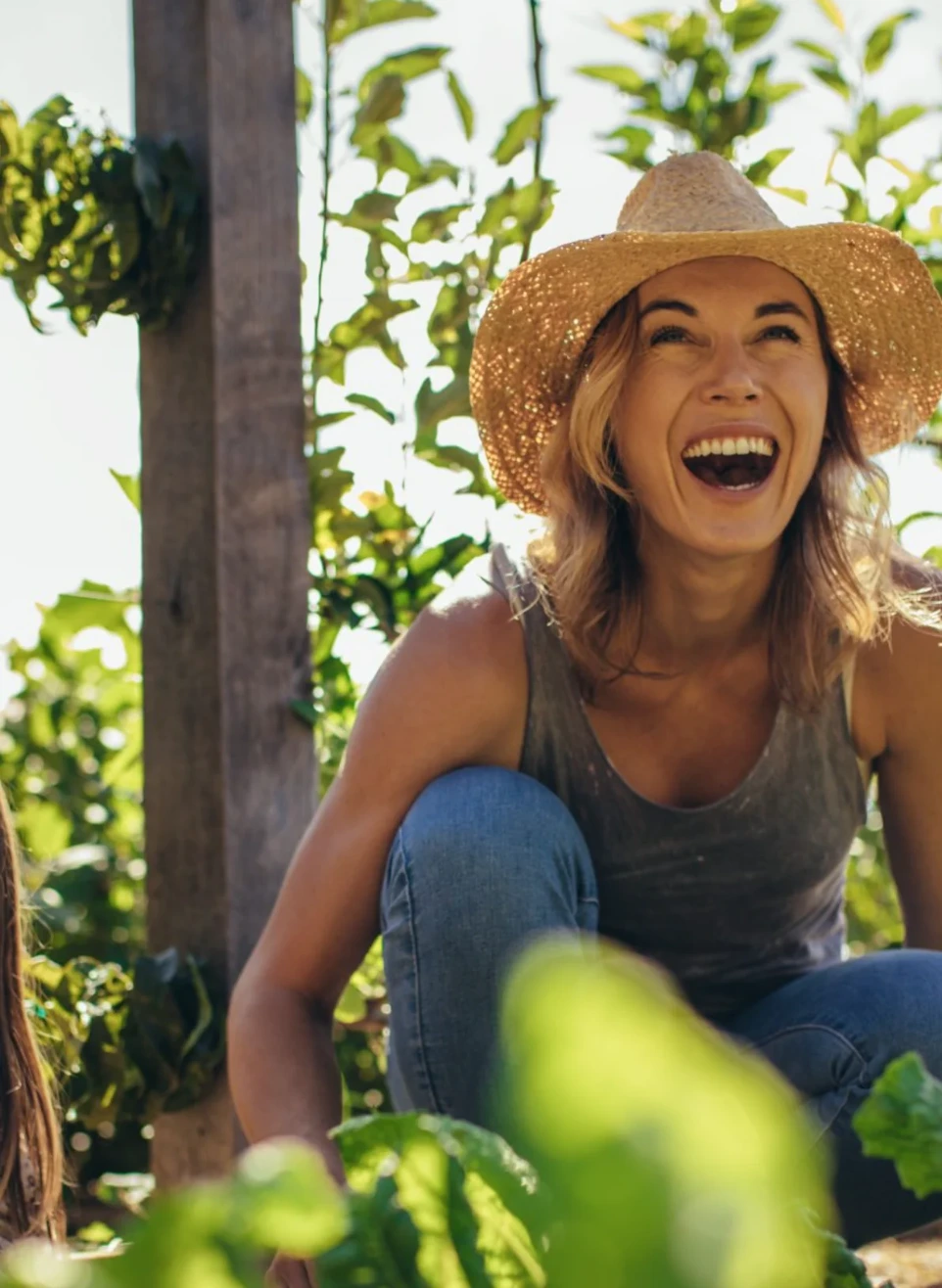 Une femme portant un chapeau de paille rit aux éclats, accroupie dans un potager ensoleillé au milieu des plantations.