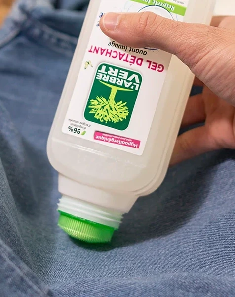 Gros plan sur l'application d'un flacon de Gel Détachant L'Arbre Vert avec son embout brosse verte directement sur un jean en toile bleue.
