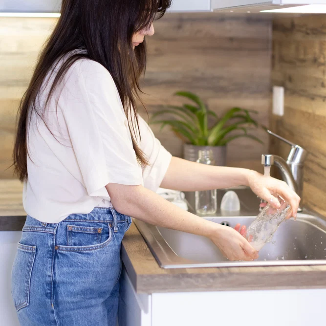 Une femme aux cheveux bruns rince un récipient en verre sous l'eau du robinet de son évier de cuisine.