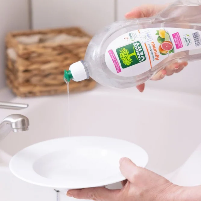 Une personne verse du liquide vaisselle main L'Arbre Vert aux Agrumes directement sur une assiette creuse blanche au-dessus d'un évier.