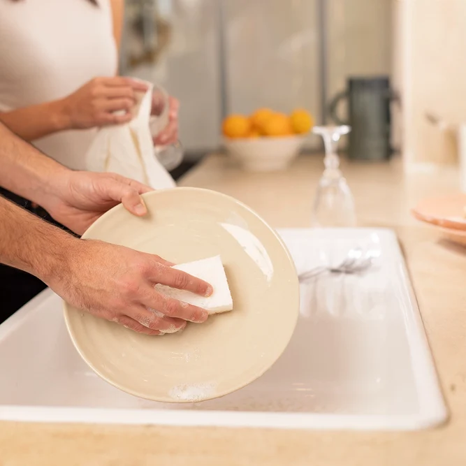 Deux mains frotte une assiette beige avec une éponge humide moussante au-dessus d'un évier, tandis qu'une personne essuie un verre au second plan.
