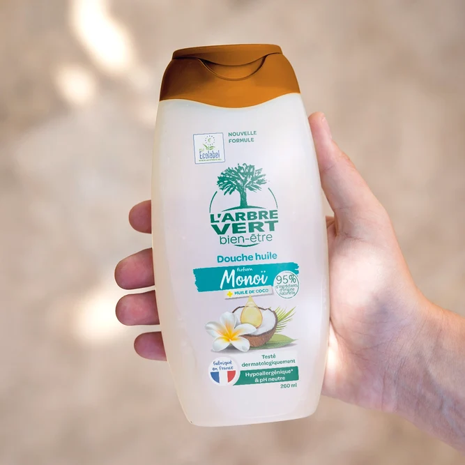 Une main tenant un flacon de 260 ml de Douche huile L'Arbre Vert bien-être au parfum Monoï et huile de coco.