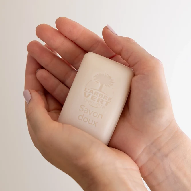 Deux mains tenant délicatement un pain de Savon doux L'Arbre Vert bien-être gravé du logo de la marque.