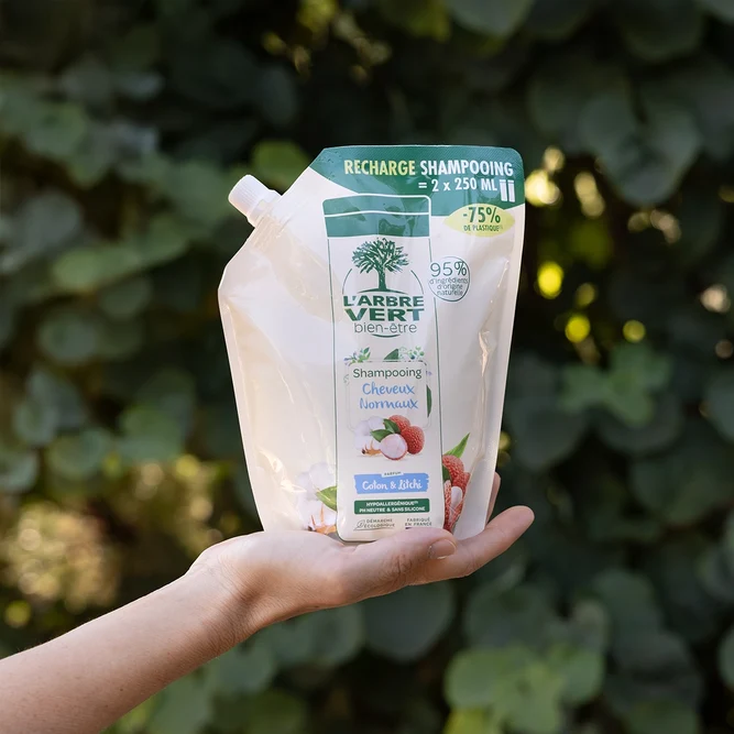 Een hand die een eco-navulling van 500 ml shampoo voor Normaal Haar L'Arbre Vert welzijn met de geur van Katoen &amp; Lychee ondersteunt, op de achtergrond van weelderig groen loof.