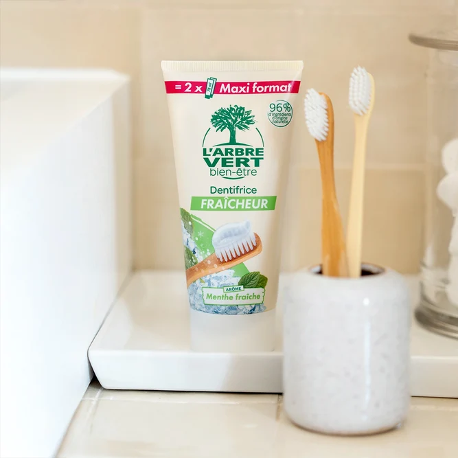 Tube de dentifrice Fraîcheur L'Arbre Vert bien-être arôme Menthe fraîche Maxi format 150 ml (équivalent à 2 tubes de 75 ml), posé sur un plateau à côté d'un gobelet contenant deux brosses à dents en bois.
