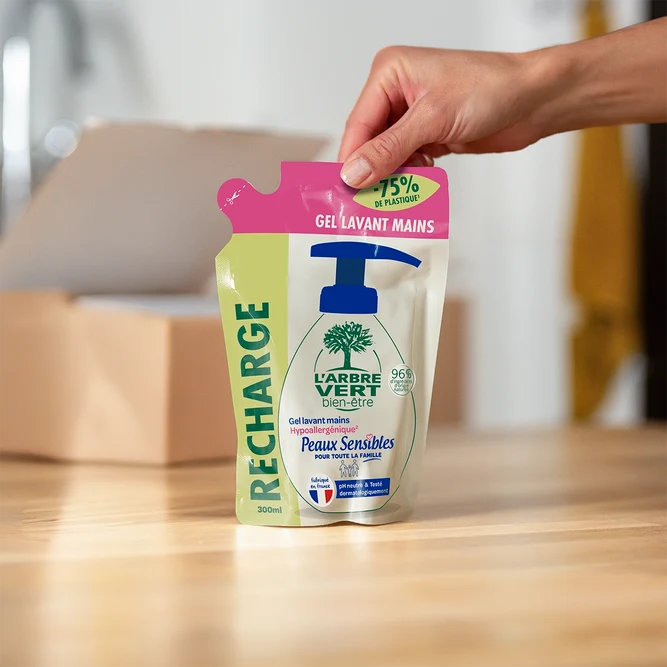 Une main posant sur une table en bois un sachet-recharge de Gel lavant mains L'Arbre Vert bien-être Peaux Sensibles de 300 ml.