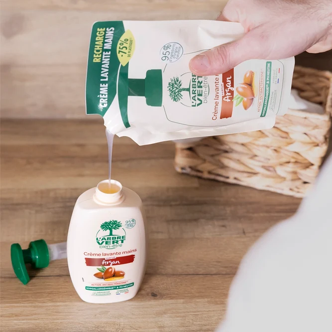 Une main versant le contenu d'un sachet-recharge de Crème lavante mains L'Arbre Vert bien-être parfum Argan (300 ml) dans un flacon dont la pompe verte a été retirée et posée sur une table en bois.